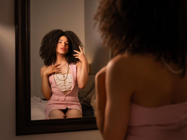 Kataleya Fowler, con i capelli ricci, un vestito rosa e un corpetto bianco con volant, si guarda allo specchio, con una mano che tocca il petto. Il riflesso mostra il suo seno e il vestito rosa..