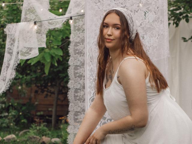 CarolinaSkye com cabelos castanhos longos, faixa de renda branca na cabeça, vestido branco, seios pequenos, tatuagem no ombro esquerdo, luzes de cordão, tecido de renda, fundo com folha verde, sorriso suave, luz natural..