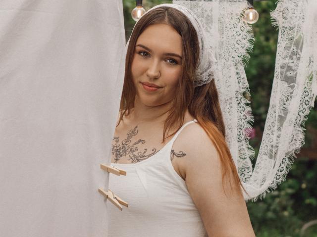 CarolinaSkye, com cabelo castanho claro, uma faixa de renda branca na cabeça, uma camiseta branca com botões de madeira e tatuagens no peito, sorrindo levemente, em pé perto de uma cortina de renda, com um fundo verde..
