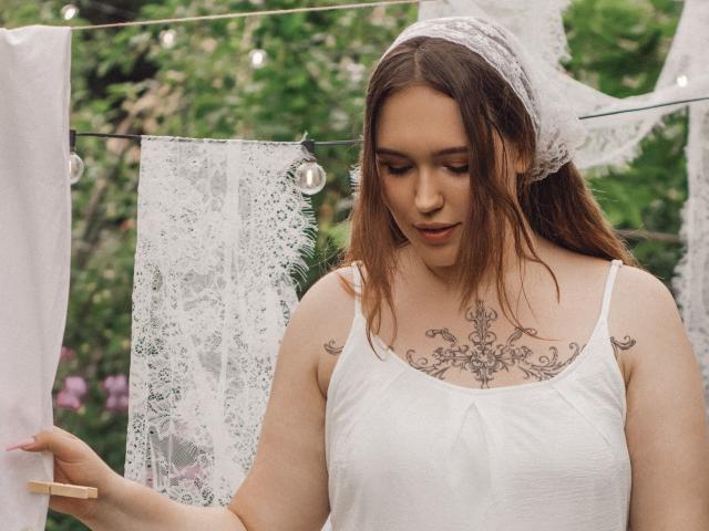 CarolinaSkye, com cabelo castanho, lenço de renda branco na cabeça, top branco e tatuagem intrincada no peito, segurando um prendedor de roupa, olhando para baixo, em um ambiente externo, com fundo verde, com roupas de renda e brancas penduradas..