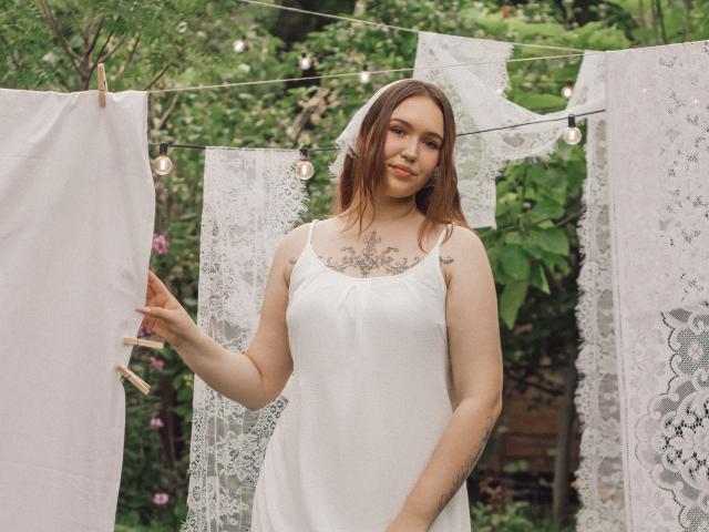 CarolinaSkye, com cabelo castanho, vestido de renda branca e tatuagens no peito, está ao lado de um varal com renda e tecido branco, segurando um pedaço de tecido com prendedores de madeira..