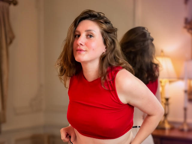 KatyaRogersX with wavy brown hair, red crop top, and white pants, smiling slightly, mid-motion, showing her tits, in a warmly lit room with a lamp in the background.