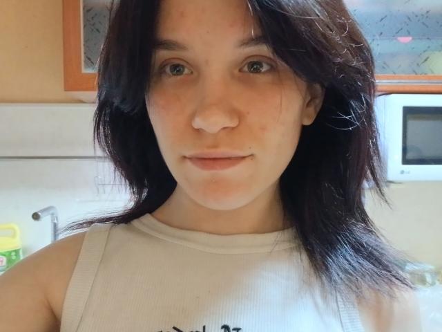 CherWinger has pale skin, dark brown shoulder-length hair, and a neutral expression. She's wearing a white ribbed tank top, showing small, subtle tits. She looks slightly flushed.