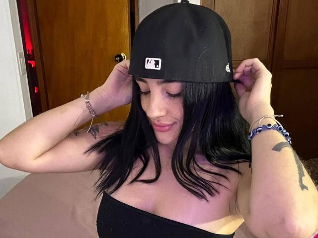 Katie Kingston mit schwarzen Haaren, einer schwarzen Mütze und einem schwarzen Top ohne Träger, das ihre Brüste zeigt, lächelnd, mit den Händen in der Mütze, Tätowierungen am Arm, Armband und Perlenarmband..