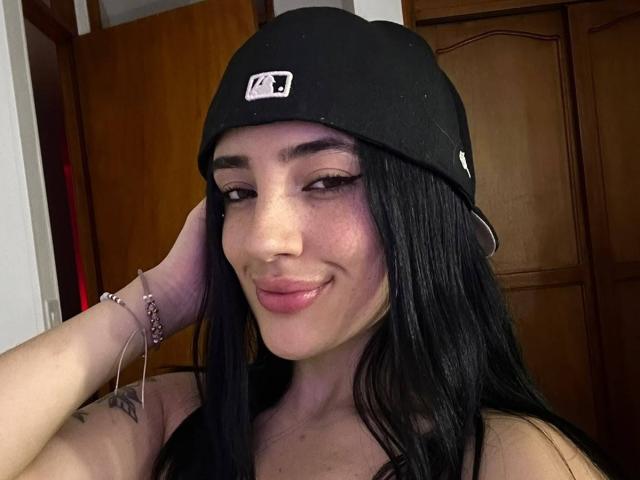 Katie Kingston, met lang zwart haar, draagt een zwarte muts, glimlacht naar de camera, heeft haar hand op haar hoofd, een roze armband om haar pols, subtiele tatoeages op haar arm en roze lippenstift..