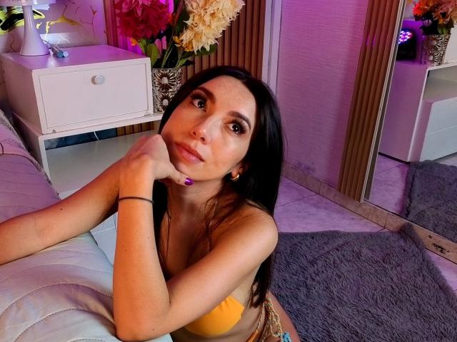 Mia Reed, con lunghi capelli neri, un top da bikini giallo e smalto per unghie viola, appoggia il mento sulla mano, mostrando il seno e i glutei piccoli, in una stanza illuminata di rosa..