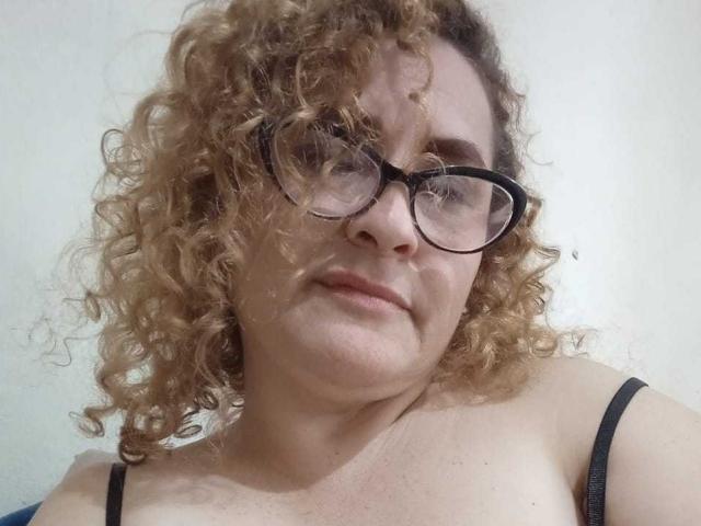 SophiiaMonroe, com cabelos cacheados, óculos, seios pequenos e um leve sorriso, vestindo uma blusa preta com alças, em um fundo simples..