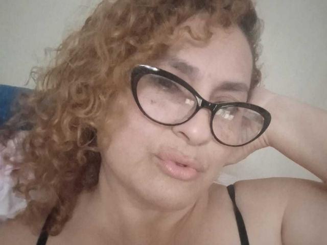 SophiiaMonroe mit lockigem, braunem Haar, schwarzen Brillen und vollen Lippen, trägt ein schwarzes Top mit Spaghetti-Trägern, legt ihren Kopf in die Hand und sieht süß und leicht genervt aus..