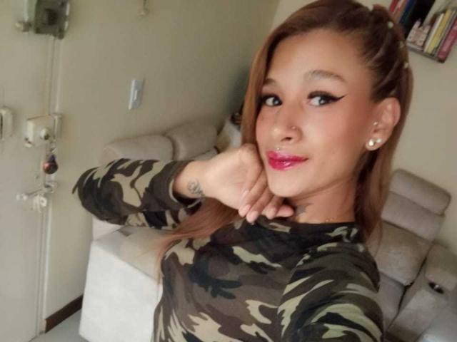 Liah Valencia, avec du rouge à lèvres rose, un anneau au nez et des piercings aux oreilles, portant un haut camouflage, tenant son menton, souriant, avec des cheveux bruns, un tatouage au cou et un petit tatouage sur la main..