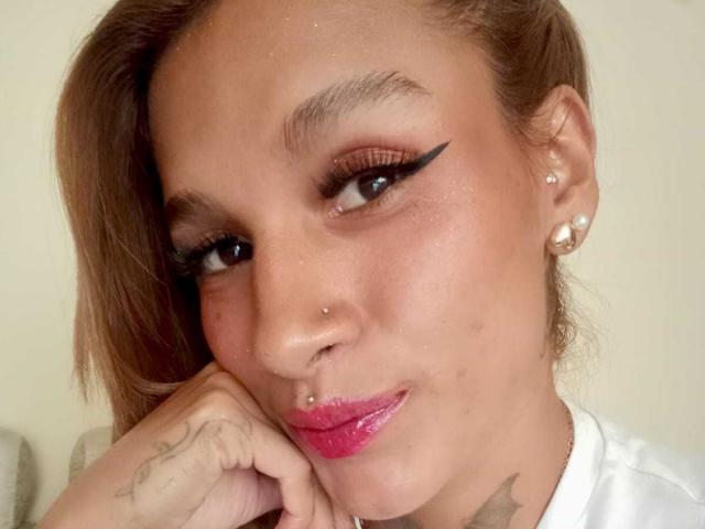 Liah Valencia, con rossetto rosa, eyeliner nero ad ali di farfalla e orecchini di perle, appoggia il mento sulla mano, mostrando un piercing al naso e un tatuaggio sul dito..