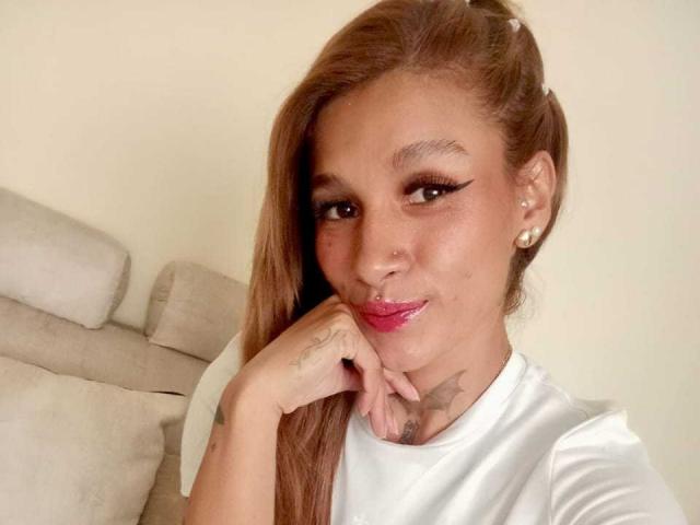 Liah Valencia, avec de longs cheveux bruns, une chemise blanche, un anneau au nez, un anneau aux lèvres, du rouge à lèvres rose, du crayon pour les yeux, des tatouages sur le cou et la main, une main sur le menton, souriante, des boucles d'oreilles en perles, peau claire..