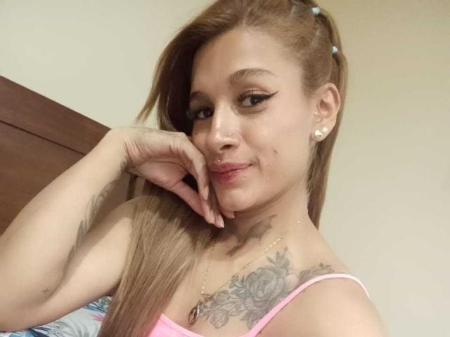 Liah Valencia, con lunghi capelli castani, una maglietta rosa, tatuaggi sul petto e un piccolo piercing al naso, sorride con una mano sul viso, indossando orecchini di perle..