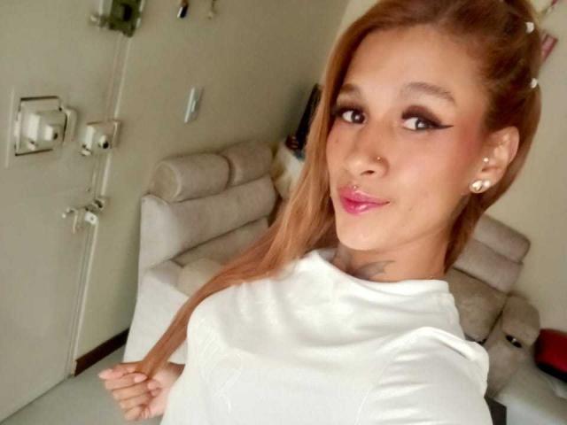 LiahValencia, avec ses longs cheveux lisses et brun-roux, son t-shirt blanc, son anneau au nez, son rouge à lèvres rose et son tatouage au cou, sourit à l'appareil photo, montrant ses seins..