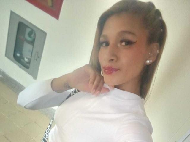 Liah Valencia, avec des lèvres roses brillantes, une chemise blanche, une main sur le cou, des cheveux bruns, des boucles d'oreilles et un tatouage au poignet, regardant l'appareil photo avec un léger sourire..