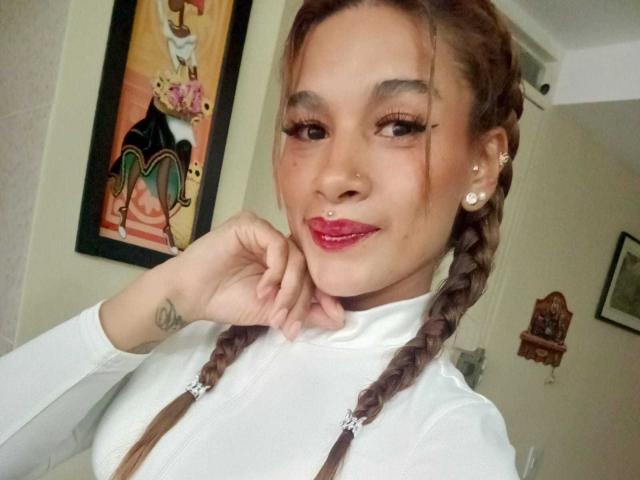 Liah Valencia, con i capelli castano intrecciati, un maglione a collo alto bianco, rossetto rosa, un anello al naso, orecchini di perle, una mano sul mento, un piccolo tatuaggio sul polso, sorride alla fotocamera..