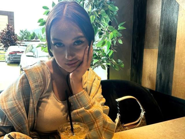 Anika Trentz, con cabello castaño corto, mano en la mejilla, camisa de cuadros, camiseta blanca, mirando a la cámara, sosteniendo una bebida con pajita, en un café acogedor, con una planta verde en el fondo..