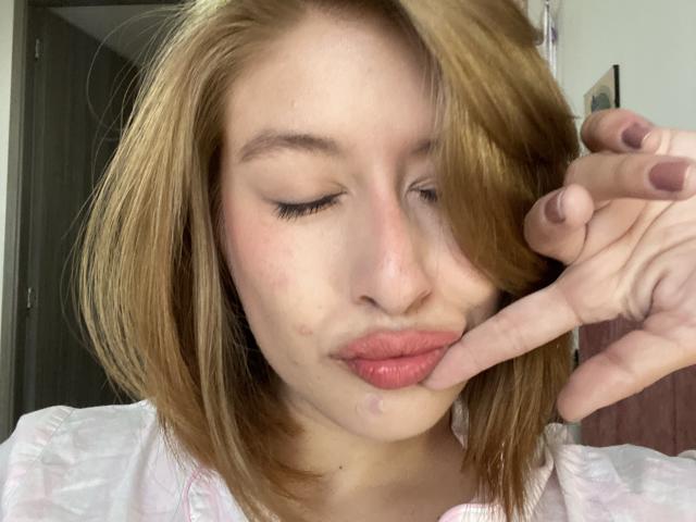 Isabella Palacio, met gesloten ogen, roze lippen en bruin haar, raakt haar lippen zachtjes aan met haar rechterhand, en draagt een witte blouse. Haar nagels zijn donkerroze geverfd..