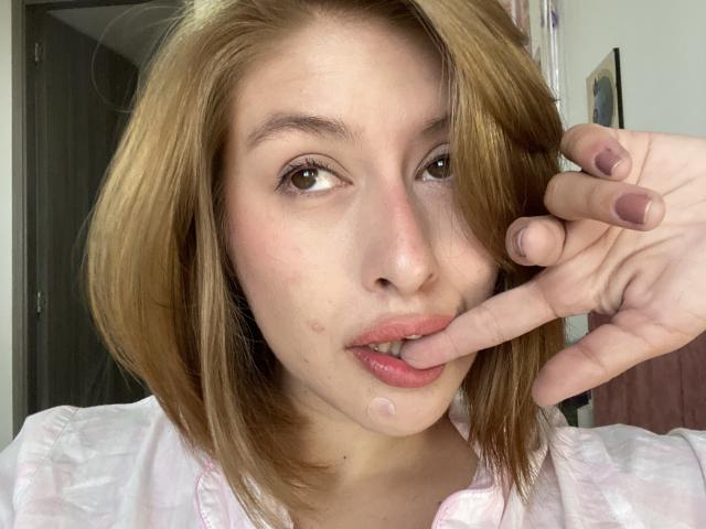 Isabella Palacio, met lichtbruin haar, die haar vinger bijt met roze lippen, draagt een witte blouse, heeft een kleine puistje op haar kin en heeft roze nagellak..