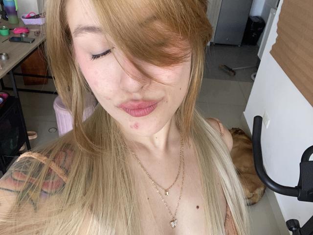 IsabellaPalacio with blonde hair, pouty lips, small red mark on cheek, gold necklace, and tattoo on shoulder, eyes closed, taking selfie, cat in background, messy room.