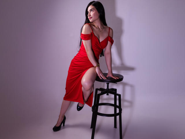 JadeHarpper, with long black hair, wearing a red off-shoulder dress, black high heels, and gold bracelet, sitting on a black stool, showing her left thigh and butt.