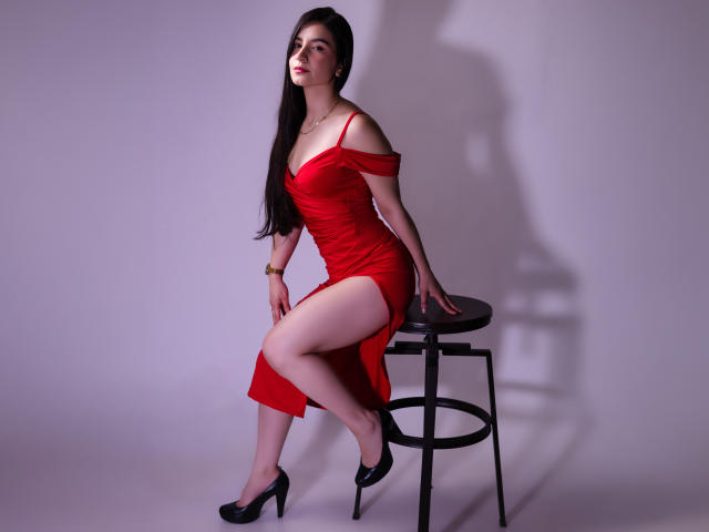 Jade Harpper, avec de longs cheveux noirs, une robe rouge à une épaule, une fente haute, des talons noirs, assise sur un tabouret noir, peau pâle, regardant sur le côté, ombre sur le mur..