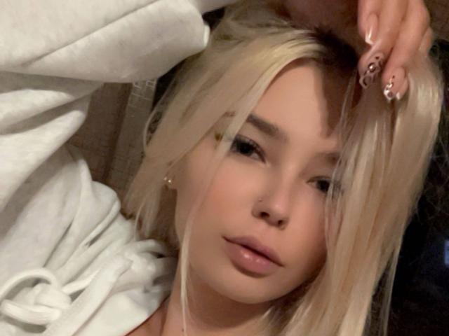 Adriene Degennaro mit blonden Haaren, leicht geöffneten Lippen, trägt eine weiße Kapuzenjacke, eine Hand im Haar, zeigt lange, gepflegte Nägel, blickt in die Kamera, weiches Licht, entspannte Atmosphäre..