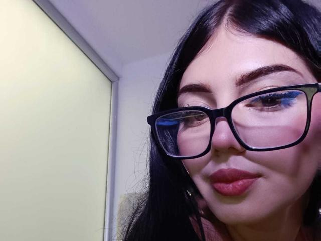 Isabella Rossi heeft donker haar, zwarte brillen, blauwe oogmake-up en rode lippenstift. Haar gezicht is lichtelijk gekanteld met een subtiele glimlach. Ze kijkt naar links..