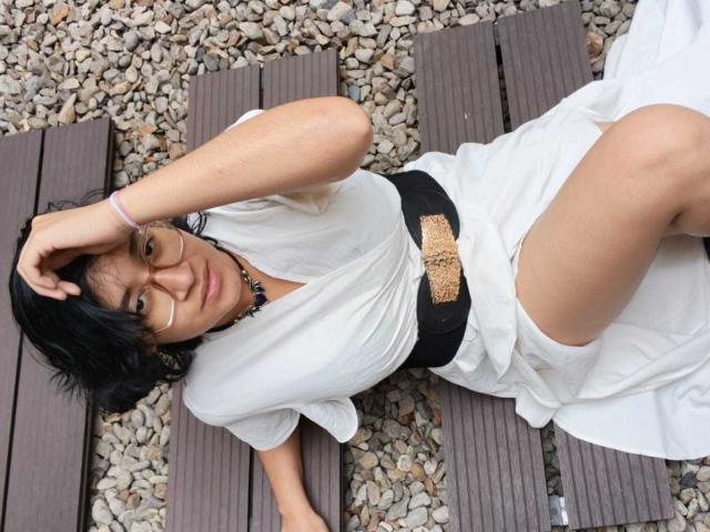 DaryMoon deitada em tábuas de madeira marrom com pedrinhas, vestindo um vestido branco, um cinto preto e um colar, tocando a cabeça com óculos, cabelo preto e uma pulseira roxa..