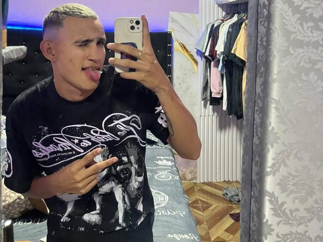 WildFantasyCouple, mit kurzen blonden Haaren, einem schwarzen T-Shirt mit Grafikmuster und der Zunge heraus, macht ein Selfie vor einem Spiegel und hält ein Handy, im Schlafzimmer, in dem Kleidung hängt..