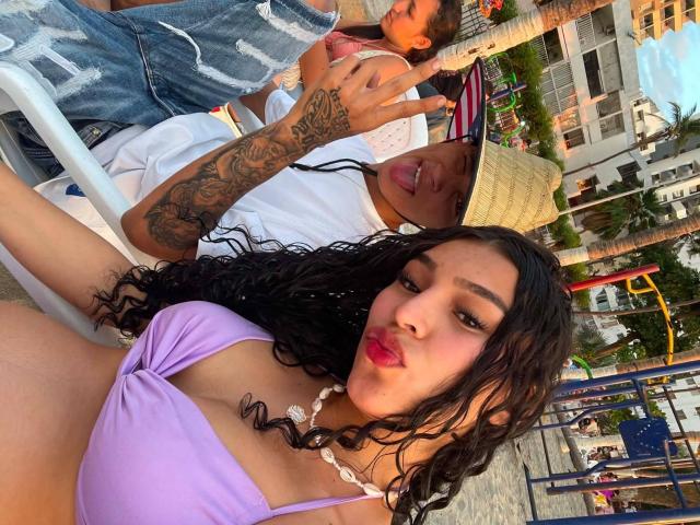 Pareja WildFantasy con cabello rizado oscuro, top de bikini púrpura, brazo tatuado y lápiz labial rosa, haciendo un gesto de paz, junto a otra pareja WildFantasy con una camisa y un sombrero blancos..