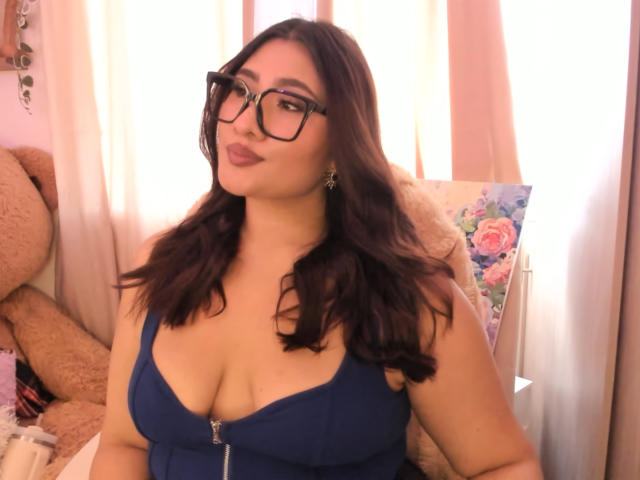 ElyRoss has large tits, wearing a blue zip-up dress, glasses, and wavy dark brown hair, with pink lipstick and a cute, confident smile in a softly lit room.