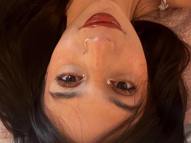 Il viso di Sophie Beaumont, con i capelli neri, gli occhi marroni, le labbra rosse lucide e una piccola goccia di liquido bianco sul naso. È sdraiata con uno sfondo rosato..