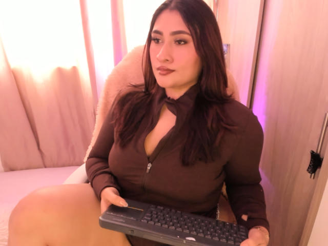 ElyRoss with brown hair, full tits in a brown zip-up, holding a keyboard, sitting in a chair, looking at the camera with a slight pout.