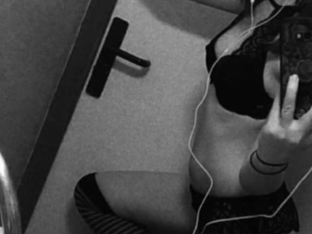CplFrancine in black lingerie, showing her tits in a lacy bra, and her striped thigh-highs, taking a selfie with white wired earbuds, in a grayscale photo.