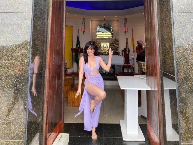 Kariina Santos, con lunghi capelli neri, indossa un vestito viola scollato con una profonda spaccatura che mostra il sedere, è in piedi in una porta, fa un gesto di pace, in una foto..