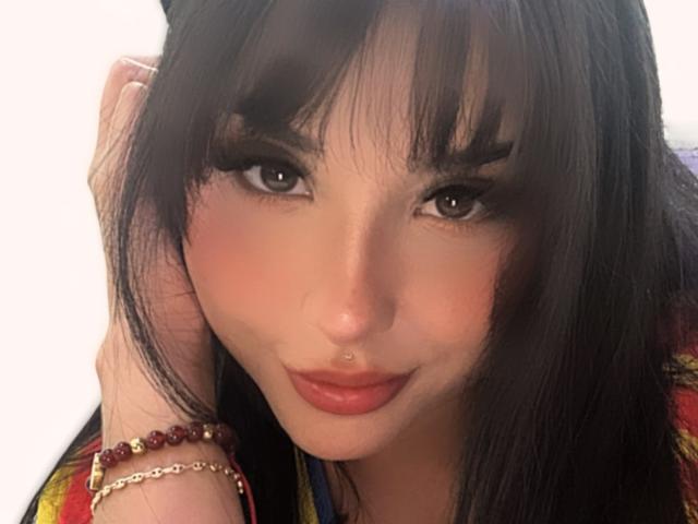 GraceClarke with black hair, bangs, and a nose ring, wearing red lipstick and a red bead bracelet, smiling subtly, brown eyes, slightly flushed cheeks, holding her hair.