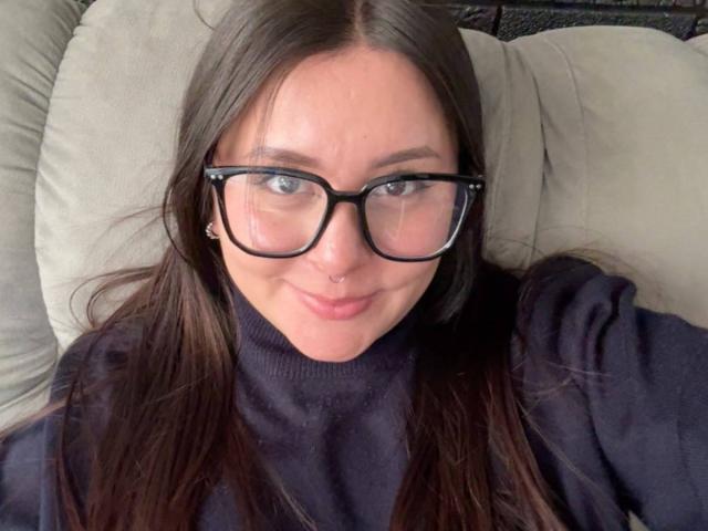 EmaJames with glasses, brown hair, nose ring, and black turtleneck, smiling slightly, showing her tits and cute face. She's lying on a beige pillow.