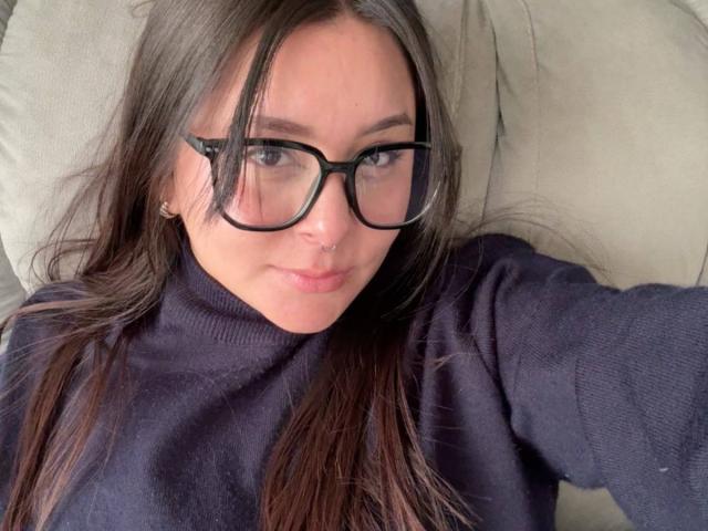 EmaJames with long brown hair, black glasses, and a nose ring, wearing a dark purple sweater, lying down and looking at the camera with a slight smile.