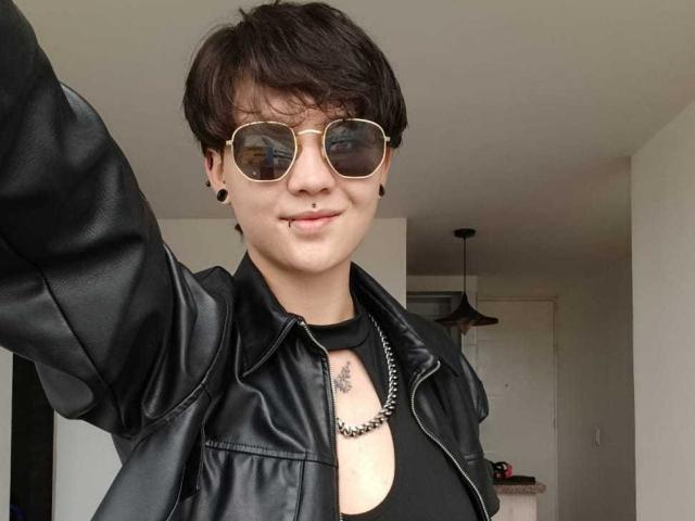 LissyLuxe with short brown hair, wearing aviator sunglasses, black leather jacket, black tank top, and black necklace, has a nose ring and lip ring, smiling. Her tits are visible.