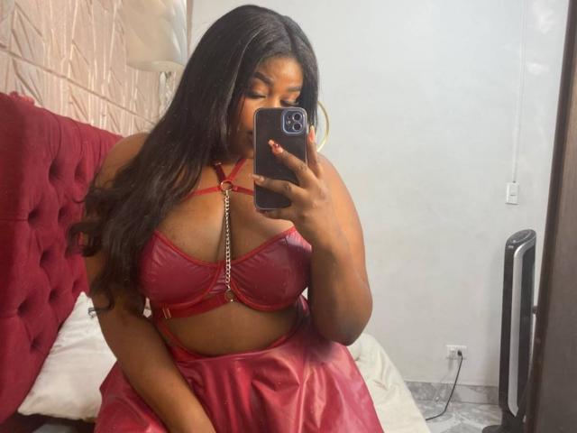 NicolZurich, con piel oscura, cabello negro largo, vestida con un arnés y falda de cuero rojo, mostrando sus grandes senos, tomando una selfie con un teléfono negro..
