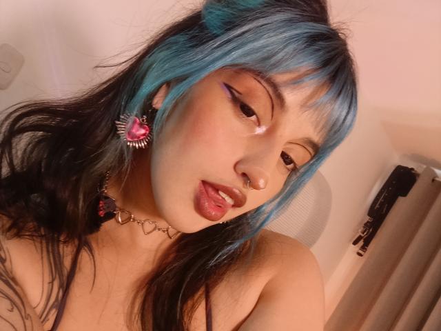 CatBleu with blue and black hair, sticking out tongue, nose ring, heart earrings, heart choker, pink eyeliner, and looking at camera. Her tits are partially visible.