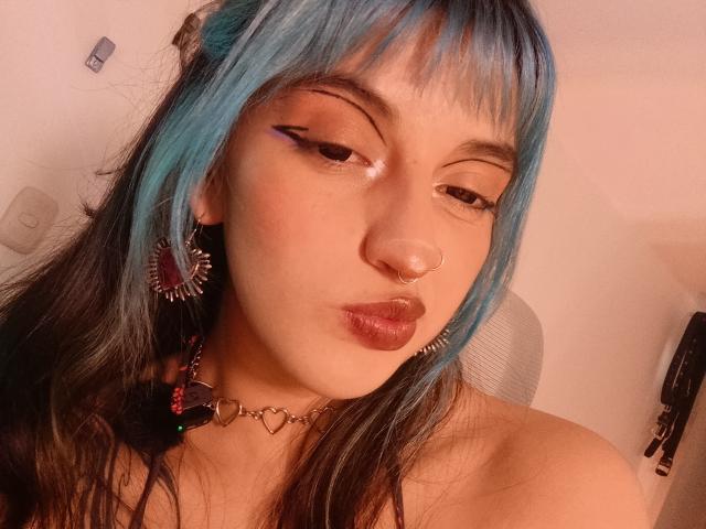 CatBleu, avec ses cheveux bleus et noirs, son anneau de nez et son fard à paupières rose, faisant la moue avec ses lèvres brillantes, portant des boucles d'oreilles à pointes et un collier en forme de cœur, dans un autoportrait éclairé par une lumière chaude..