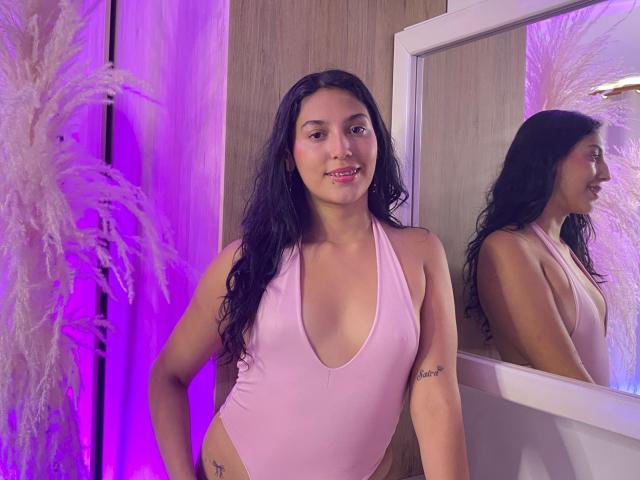 ZarethDilucas indossa un costume intero rosa, con un profondo scollo, che mette in mostra il seno e un tatuaggio sul fianco, ed è in piedi con un sorriso, riflessa in uno specchio, con illuminazione viola e piante voluminose..