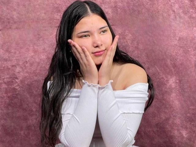 LizzieSweets, avec de longs cheveux noirs, les mains sur les joues, souriante, portant un haut blanc côtelé à une épaule, laissant légèrement entrevoir ses seins, sur un fond rose texturé..