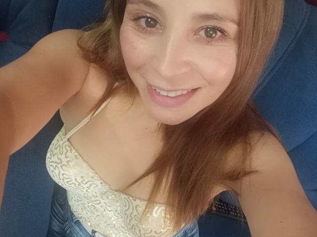 Crisht, com cabelo castanho claro, sorrindo, vestindo uma blusa branca de renda, que revela seus pequenos seios, sentada em uma cadeira azul. Seus olhos castanhos e lábios rosados são visíveis..