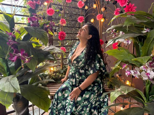 Genesis Montenegro, con cabello negro rizado, un vestido floral verde y un trasero visible, está de pie entre flores rosas y moradas, rodeada de hojas verdes y una ventana de metal ornamentada..