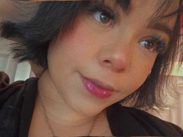 ViviWollds has medium brown skin, black shoulder-length hair, brown eyes, pink glossy lips, and a small nose ring. She's wearing a black top with a gold necklace.
