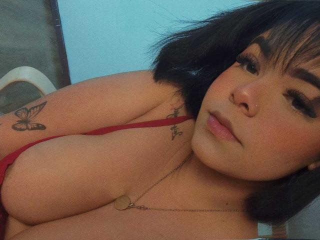 ViviWollds, con i capelli neri, indossa una maglietta rossa, ha un seno abbondante, un tatuaggio a forma di farfalla sul braccio sinistro, una collana con un cuore ed è ammiccante con un leggero sorriso..