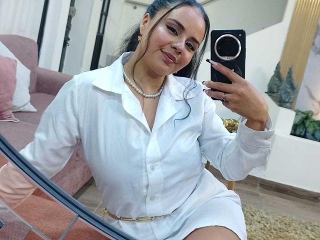 Arielle Wilde, con i capelli scuri raccolti in una coda di cavallo, indossa una camicia bianca con bottoni e una collana di perle, è seduta e sta scattando un selfie allo specchio, mostrando il suo seno..