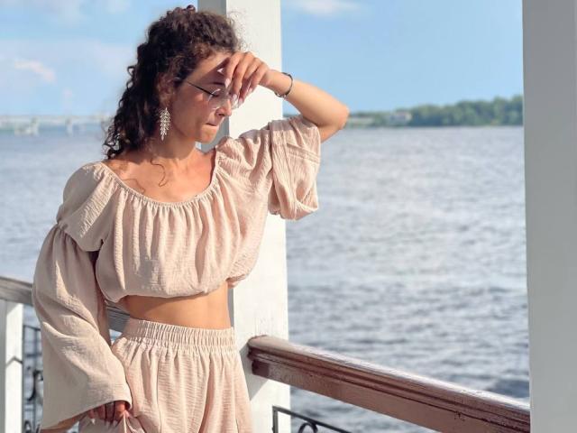 AlyceErway mit lockigem Haar, Brille und hängenden Ohrringen, trägt ein beiges, schulterfreies Crop-Top und dazu passende Hosen. Sie steht an einem Geländer und hält sich mit der Hand vor die Augen, um das Licht abzuschirmen..